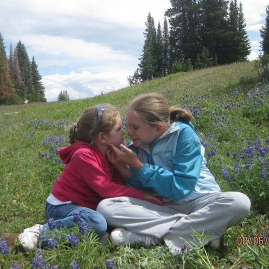 Elementary-aged sisters Jalyn and Carlin sitting in a flower-filled field, one holding the other's face closely, smiling.