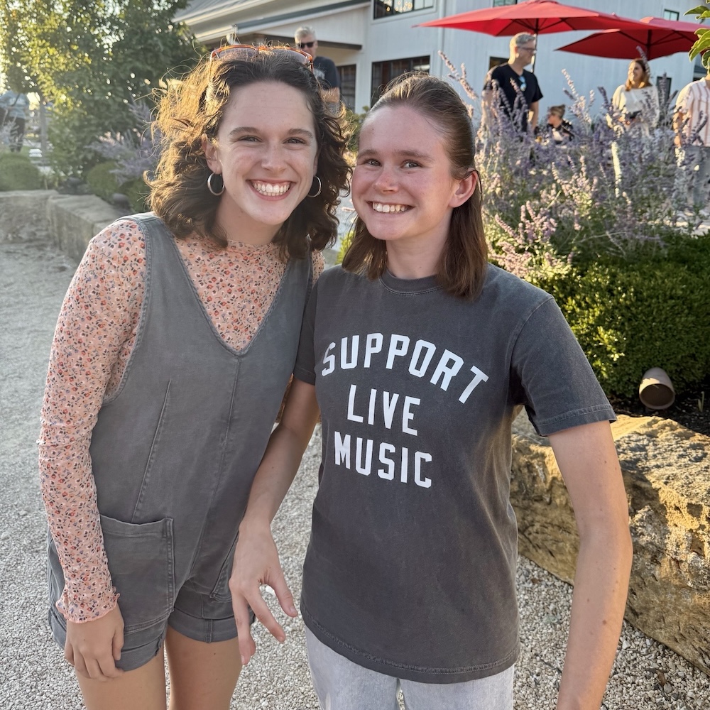 Young adult sisters Jalyn and Carlin standing in an outdoor garden venue, smiling joyfully. Carlin's shirt says 'Support Live Music'